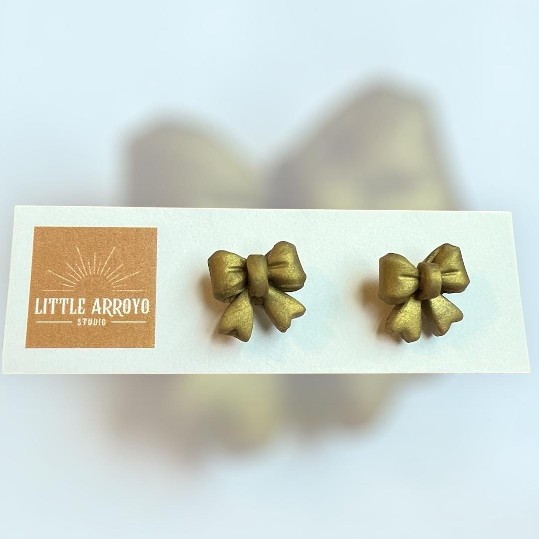 Gold delicate bow-shaped earrings on a white card in front of a blurred background. Handmade polymer clay hypoallergenic stainless steel daily everyday cute coquette femme flirty girly