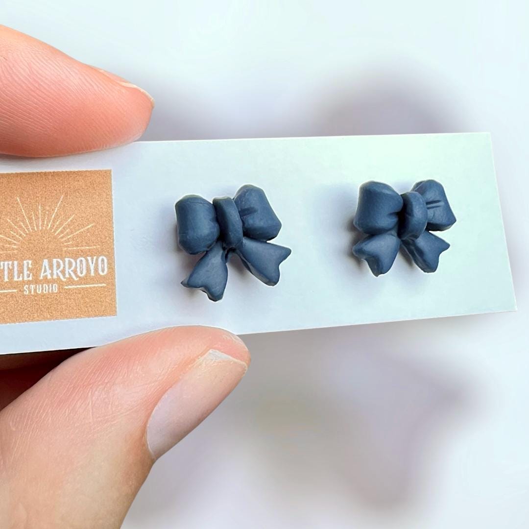 Navy blue delicate bow-shaped earrings on a white card held by a hand in front of a blurred background. Handmade polymer clay hypoallergenic stainless steel daily everyday cute coquette femme flirty girly