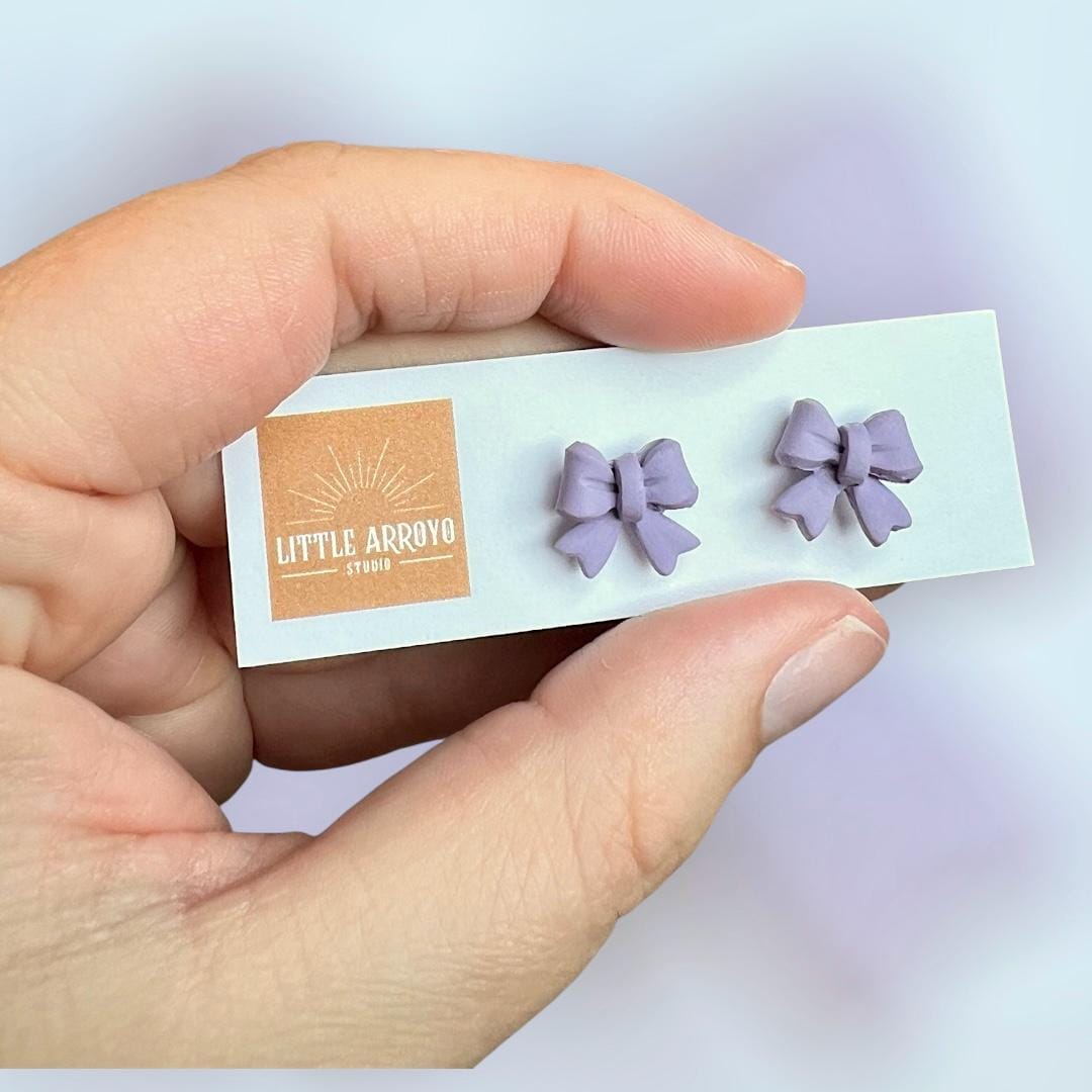Light purple, lilac delicate bow-shaped earrings on a white card held by a hand in front of a blurred backgroundl. Handmade polymer clay hypoallergenic stainless steel daily everyday cute coquette femme flirty girly