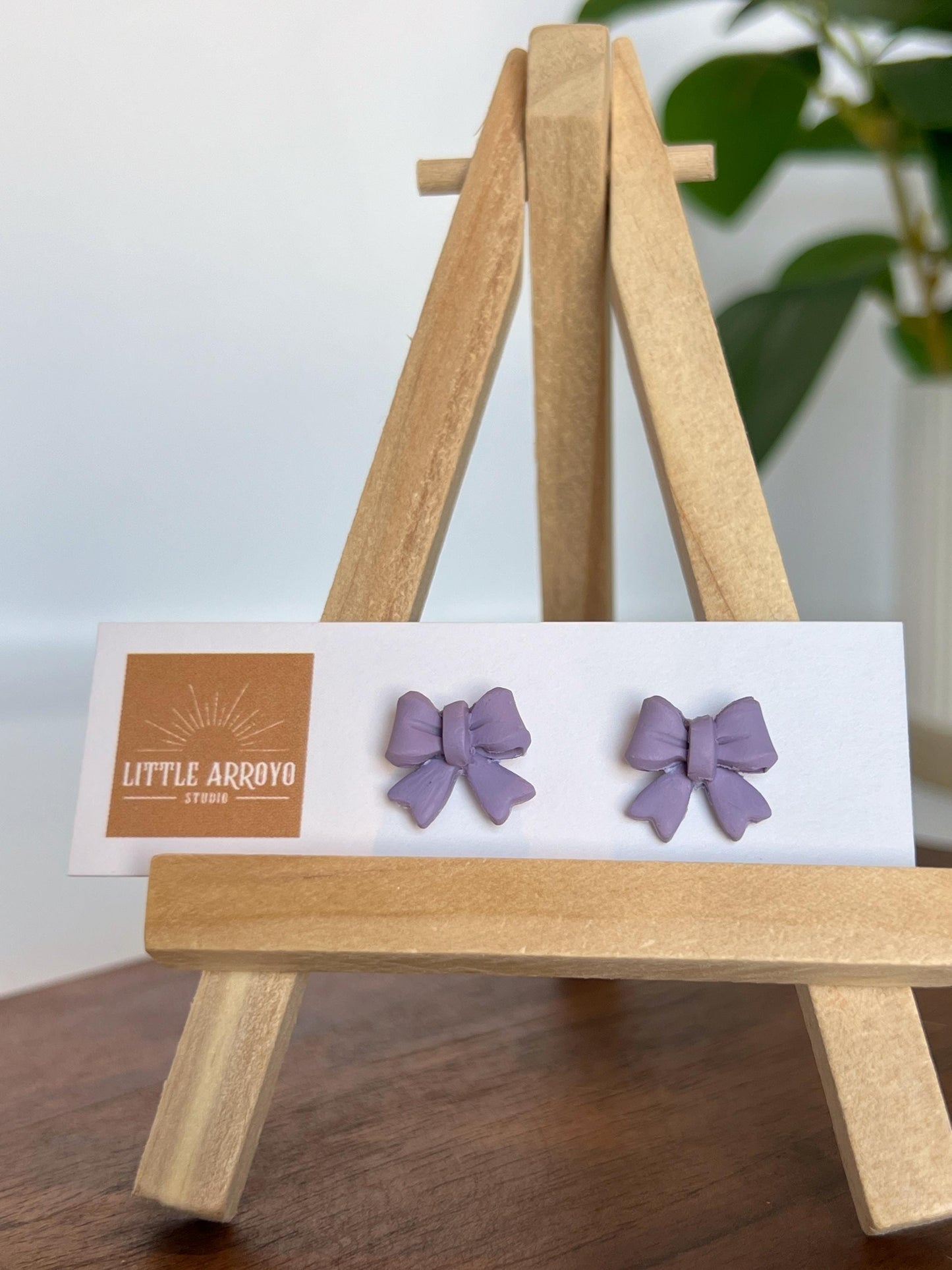 Light purple, lilac delicate bow-shaped earrings on a white card displayed on a small wooden easel. Handmade polymer clay hypoallergenic stainless steel daily everyday cute coquette femme flirty girly