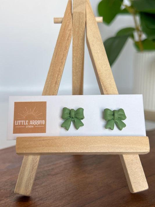 Green, delicate bow-shaped earrings on a white card displayed on a small wooden easel. Handmade polymer clay hypoallergenic stainless steel daily everyday cute coquette femme flirty girly