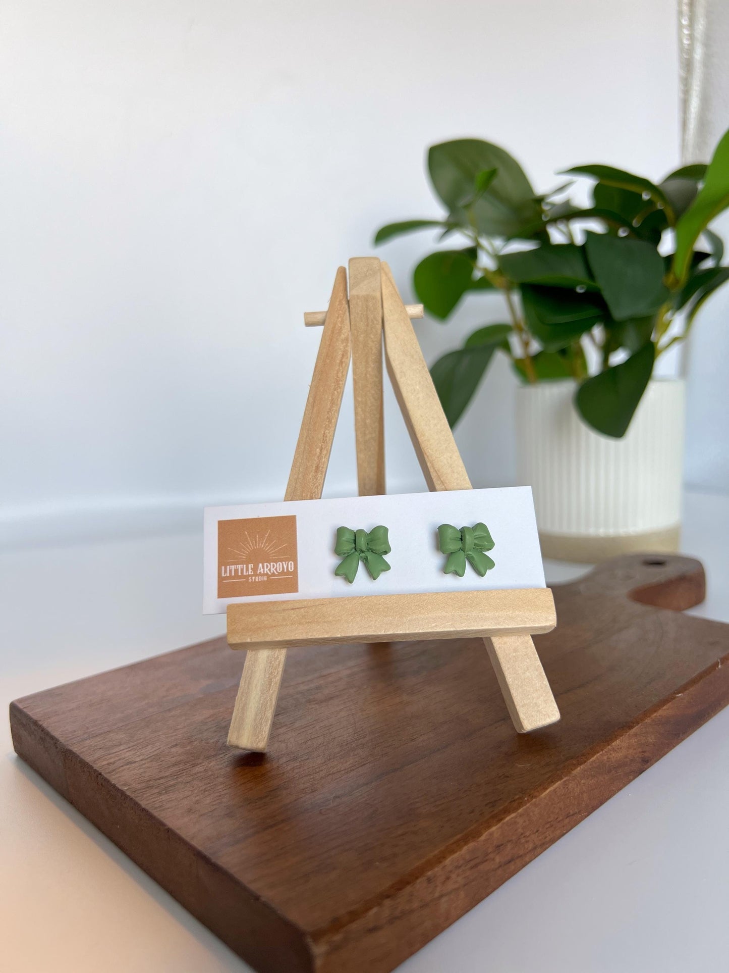 Green, delicate bow-shaped earrings on a white card displayed on a small wooden easel. Handmade polymer clay hypoallergenic stainless steel daily everyday cute coquette femme flirty girly