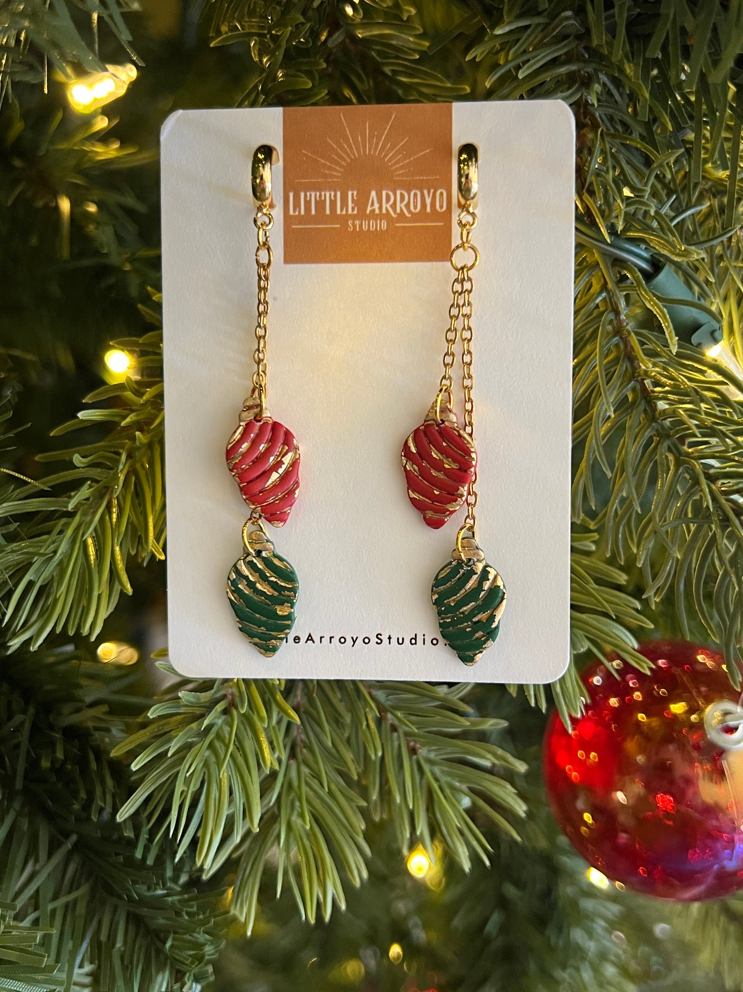 Pair of Christmas light bulb handmade polymer clay earrings on a card with Christmas tree and decorations in the background. Earrings made in Colorado by hand lightweight hypoallergenic