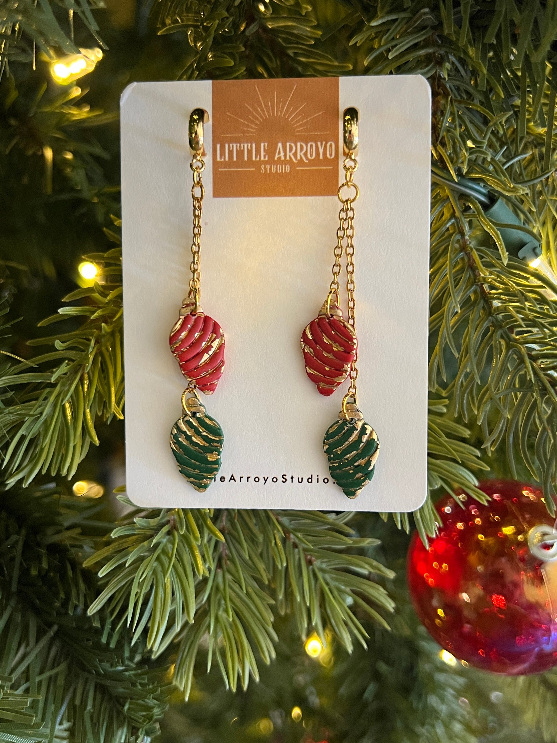 Pair of Christmas light bulb handmade polymer clay earrings on a card with Christmas tree and decorations in the background. Earrings made in Colorado by hand lightweight hypoallergenic