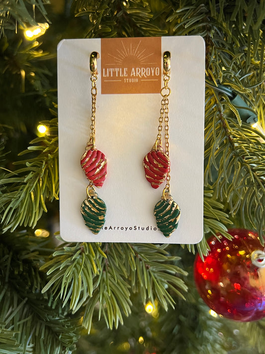 Pair of Christmas light bulb handmade polymer clay earrings on a card with Christmas tree and decorations in the background. Earrings made in Colorado by hand lightweight hypoallergenic
