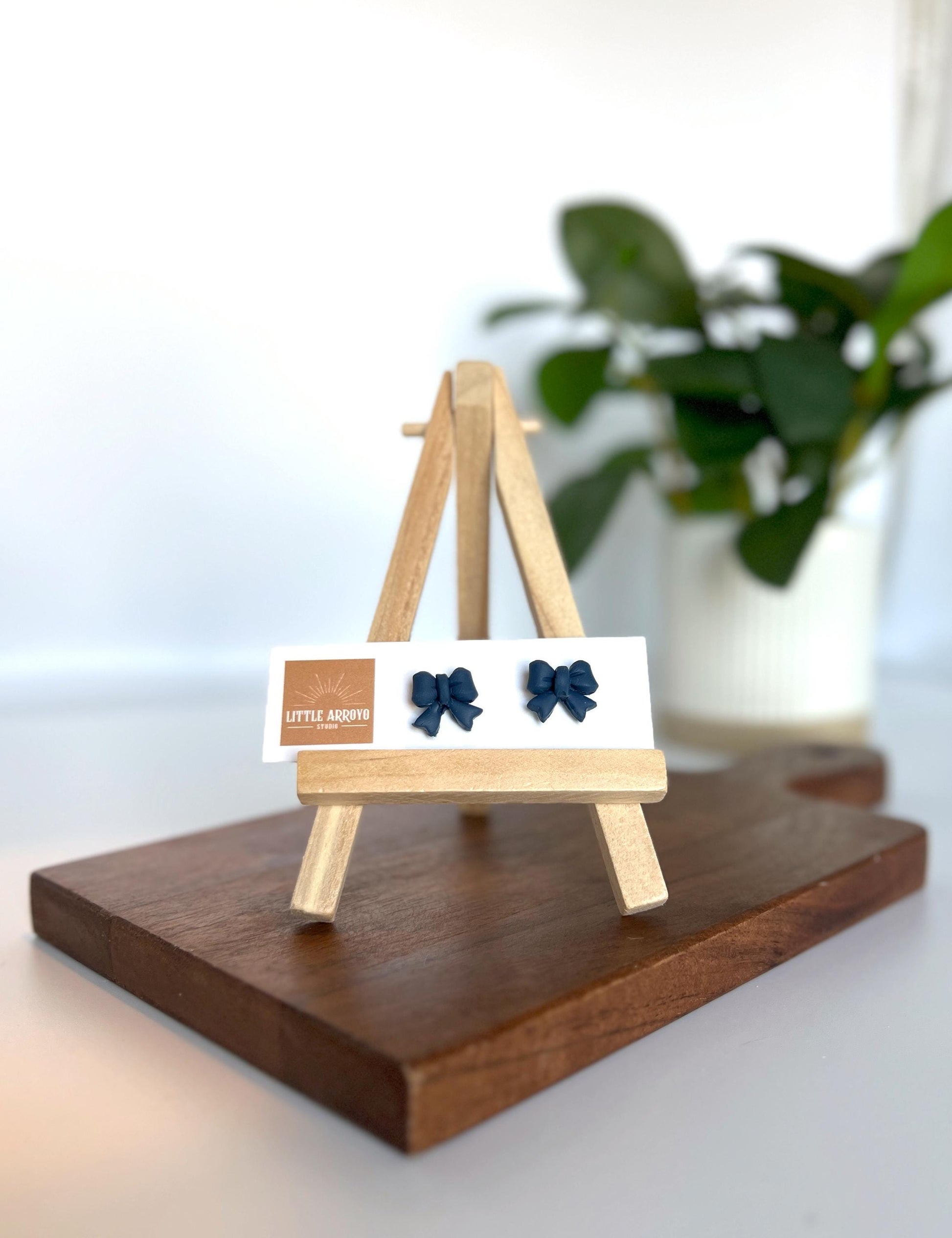 Navy blue delicate bow-shaped earrings on a white card displayed on a small wooden easel. Handmade polymer clay hypoallergenic stainless steel daily everyday cute coquette femme flirty girly