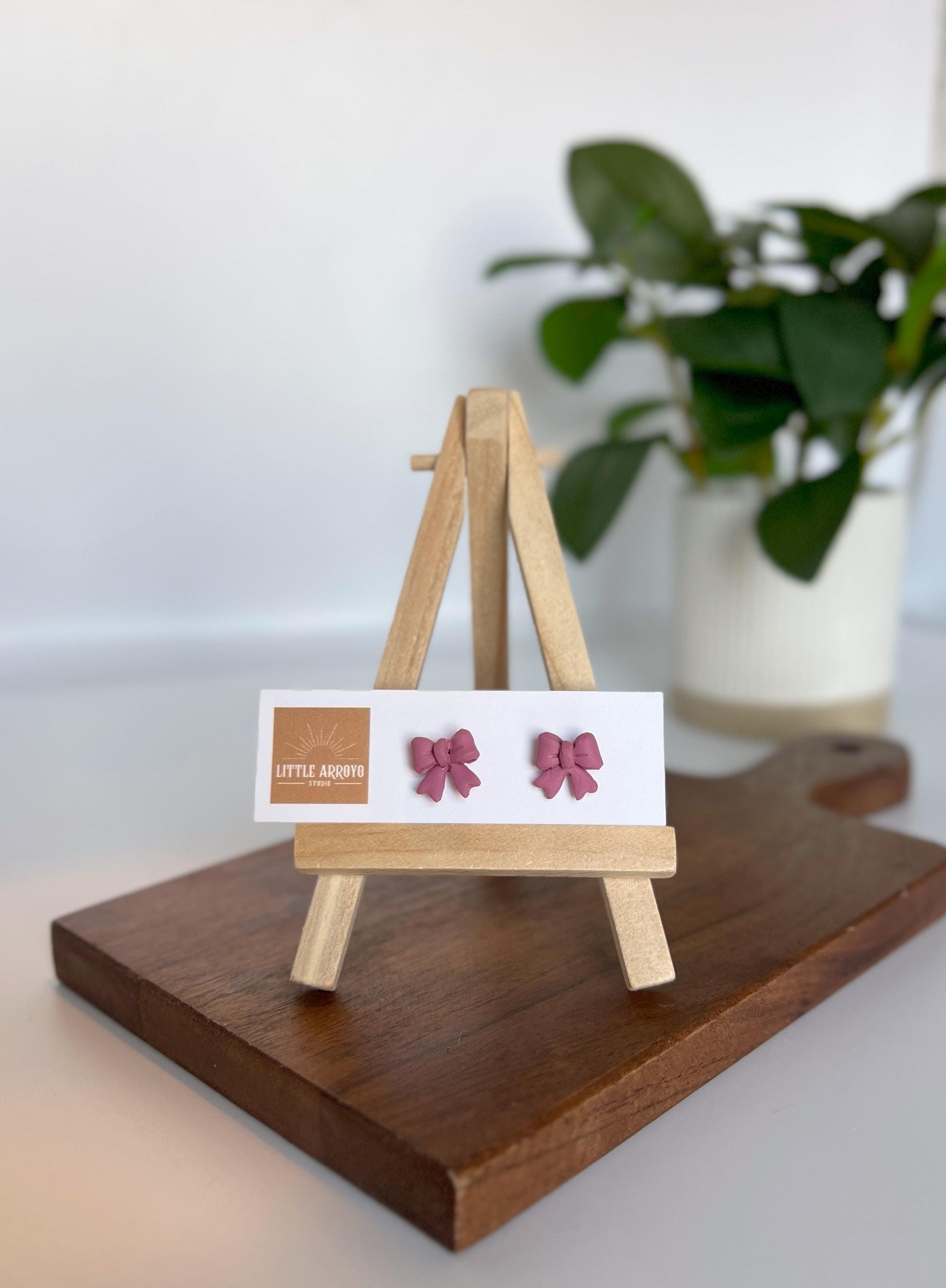 Pink, delicate bow-shaped earrings on a white card displayed on a small wooden easel. Handmade polymer clay hypoallergenic stainless steel daily everyday cute coquette femme flirty girly