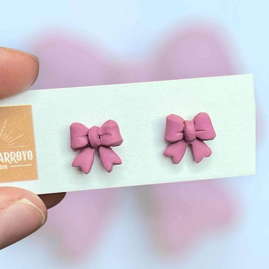 Pink, delicate bow-shaped earrings on a white card in front of a blurred background. Handmade polymer clay hypoallergenic stainless steel daily everyday cute coquette femme flirty girly