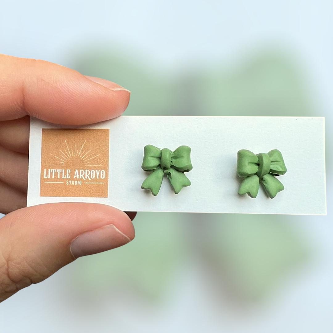 Green, delicate bow-shaped earrings on a white card held by a hand in front of a blurred background. Handmade polymer clay hypoallergenic stainless steel daily everyday cute coquette femme flirty girly